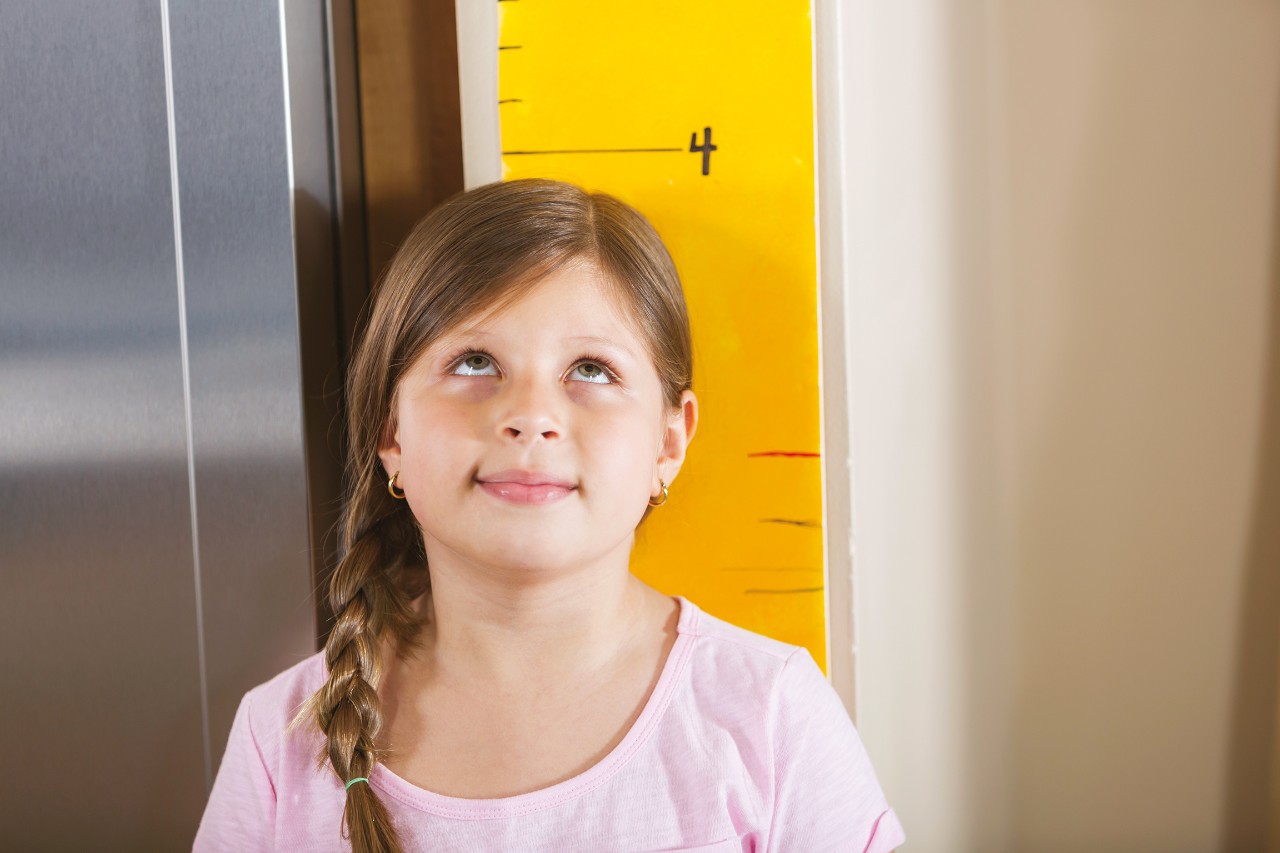 An Hispanic girl wearing a pink shirt, measuring her height, standing against a growth chart hanging on the wall at home. She is looking up with a somewhat serious expression on her face.